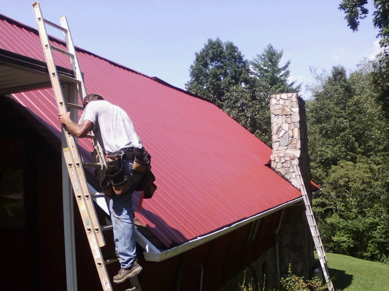 Roofer on a ladder beside a red metal roof and stone chimney — Soffit & Fascia Repair by Roofing Brothers in Richmond